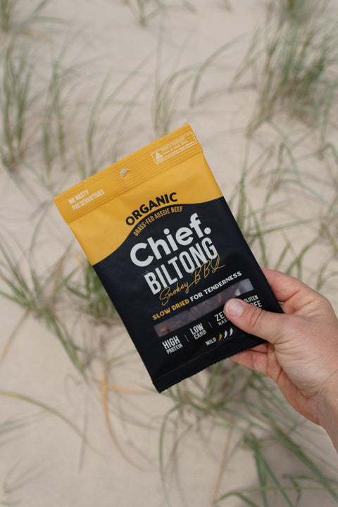 A person's hand holding a bag of Chief Smokey BBQ Biltong against a sand dune backdrop